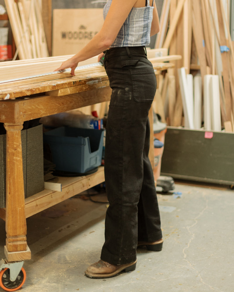Woman in Dirty Birdy Carpenter Pant in a wood shop, showcasing their functionality.